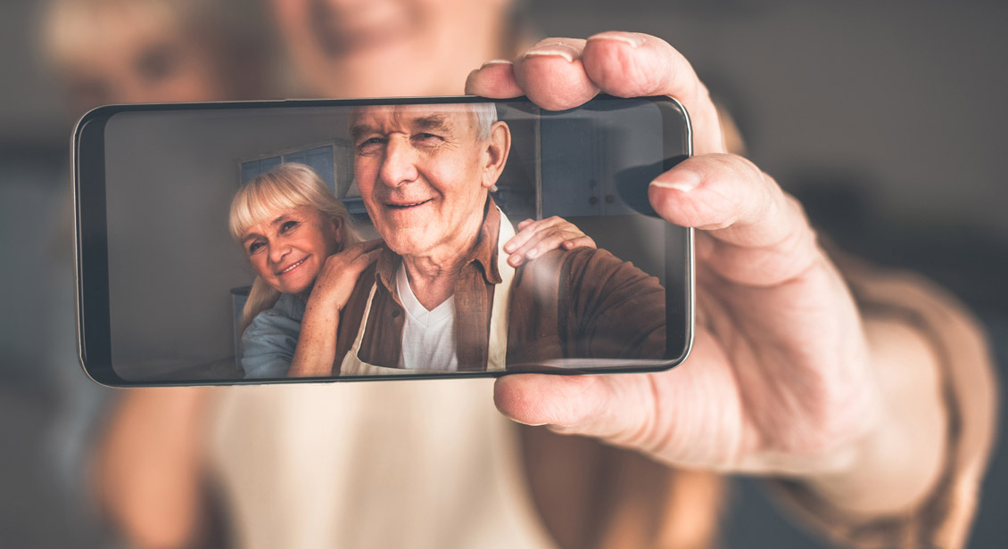 In focus phone screen held in the left hand which displays a smiling seniors couple (man and woman) on the screen of the mobile phone after taking a picture 