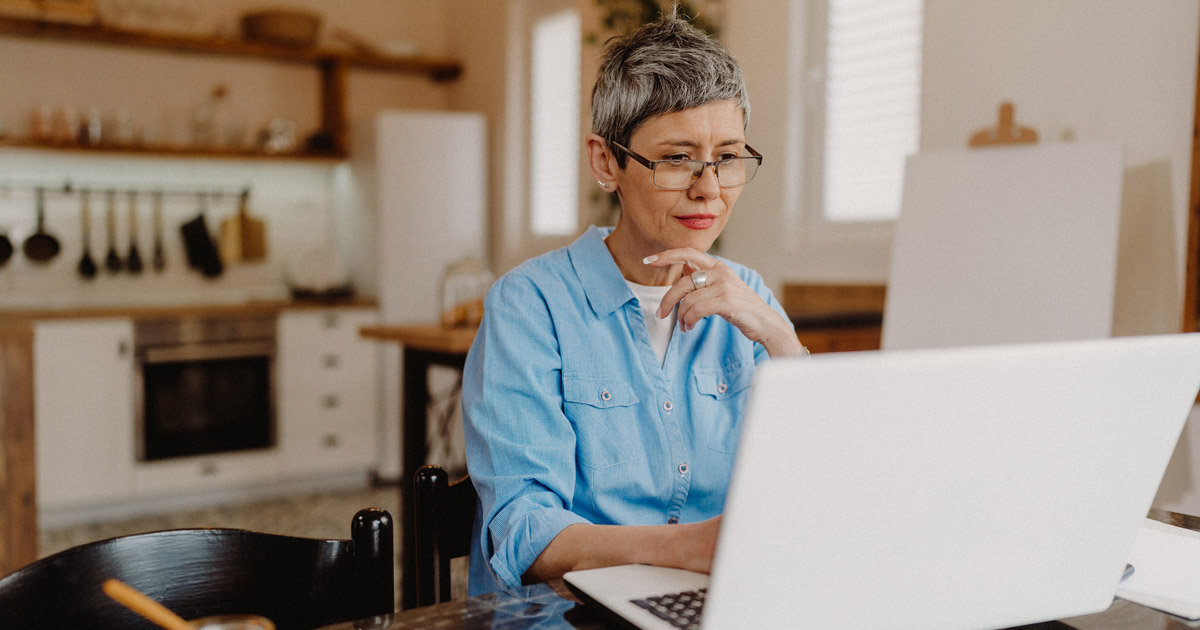 Senior aged woman with short black and grey hair sitting at her dining table using her laptop computer