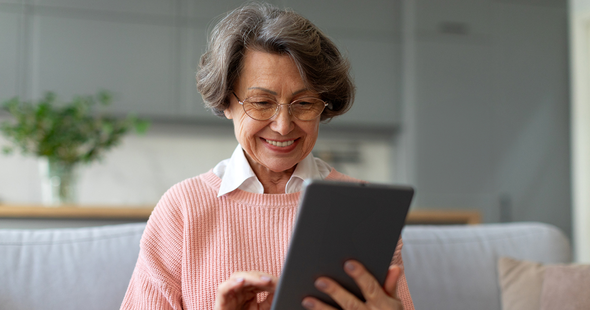 Senior aged woman in pink jump smiling and looking a tablet device held in her left hand