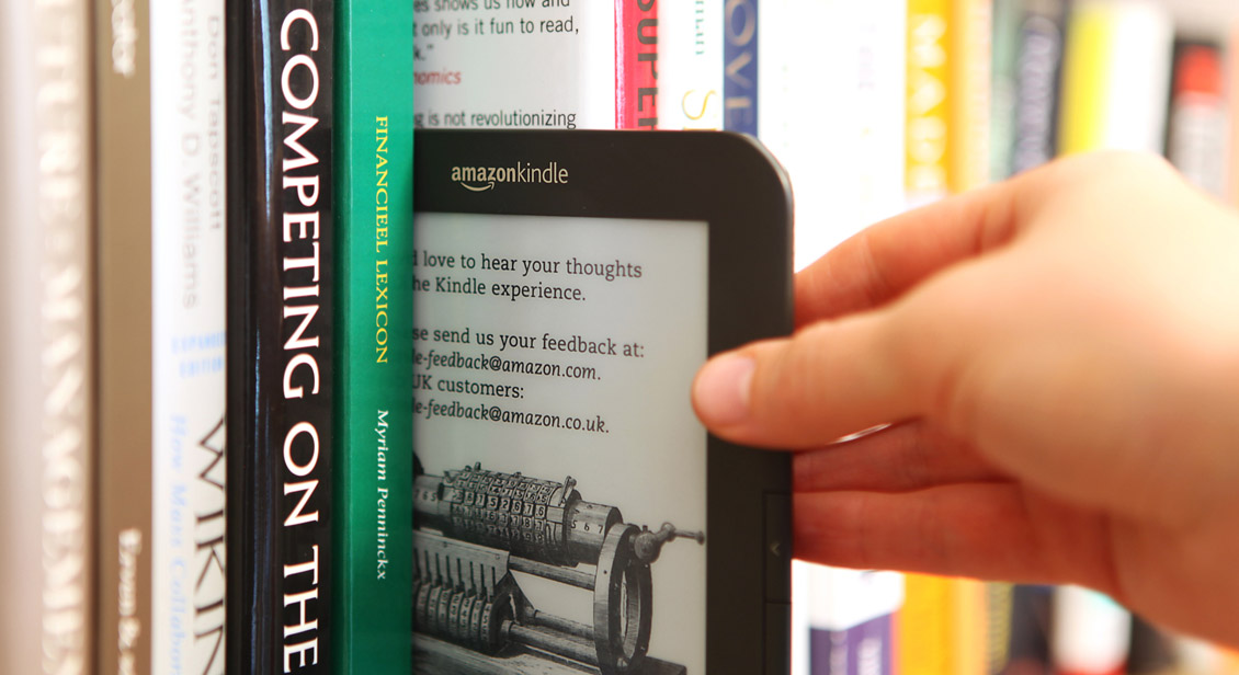 Kindle ebook reader being removed from bookshelf containing traditional paper books.