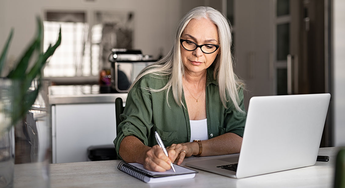 Woman with laptop writing notes on notepad.