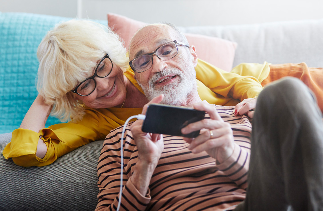 Male and female couple in their sixties relax on sofa, male shares digital content using mobile phone.