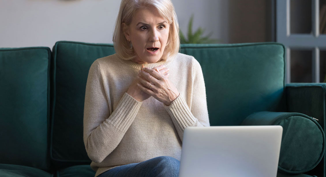 Older woman sitting on green couch with laptop looking shocked.
