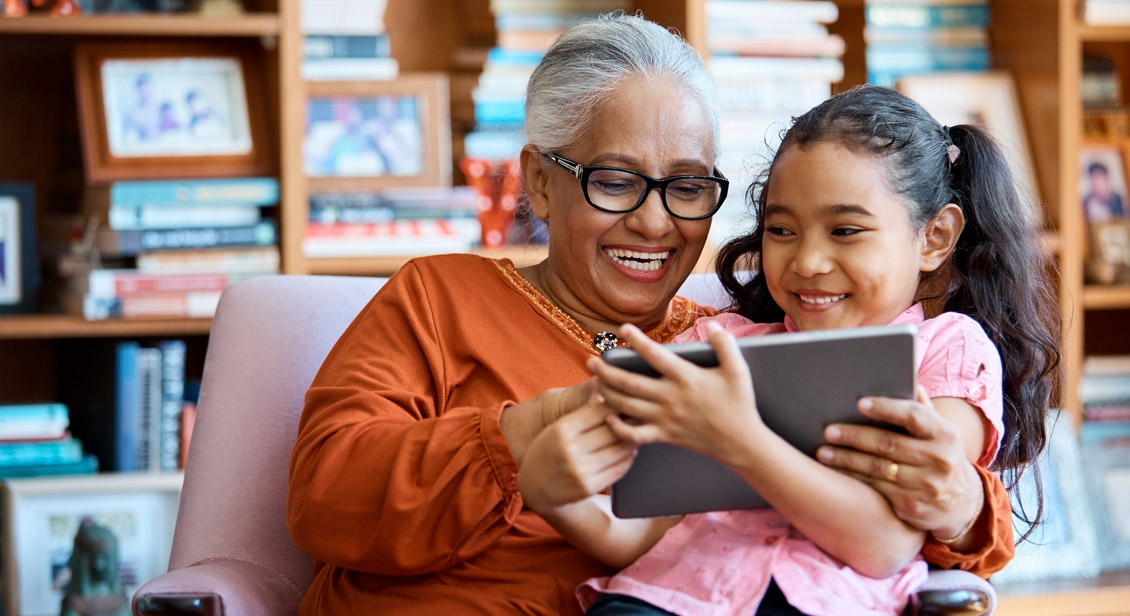 Smiling polynesian grand daughter sitting on her smiling grandmother's lap whilst sharing a tablet device