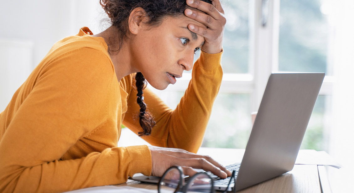 Light skinned woman with left hand on forehead looking at a laptop computer screen with a worried look on her indicating she has been scammed 
